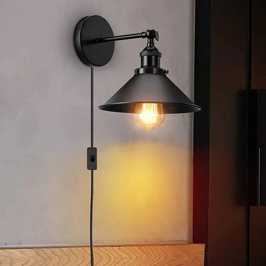 Black industrial wall lamp with plug-in cable and switch, casting warm illumination against a light neutral wall.