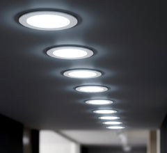 Perspective view of a straight line of circular flush recessed LED lights illuminating a dark interior hallway with cool white light.