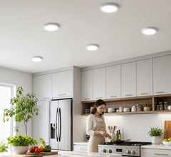 Bright, modern kitchen interior featuring round ceiling lights, light gray cabinetry, and a woman cooking at the stove.