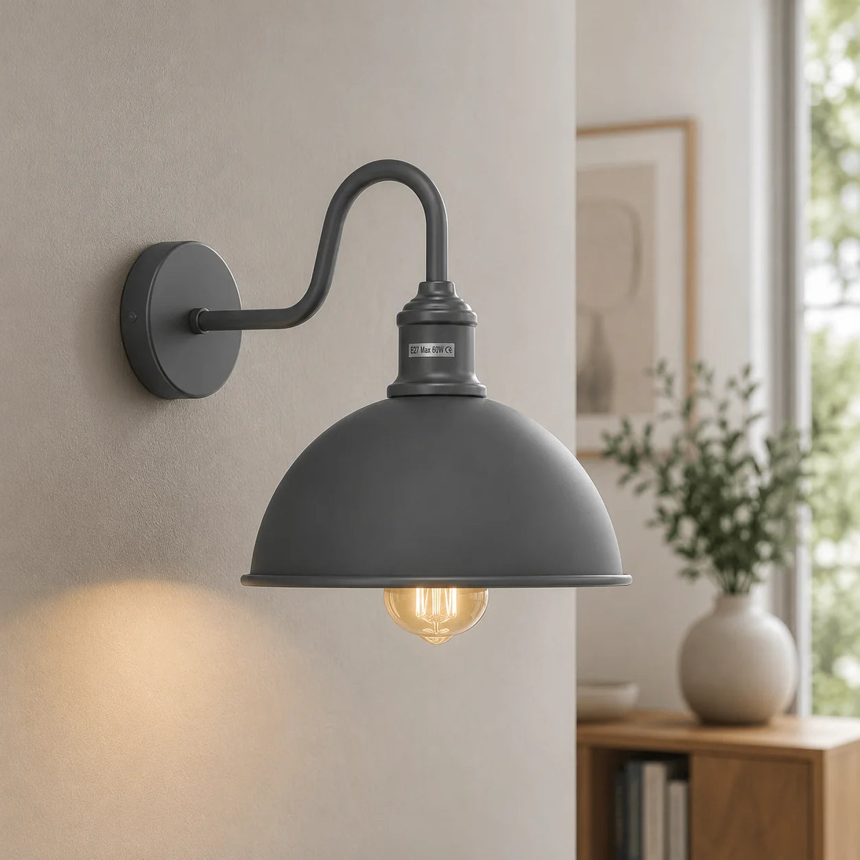 A dark gray industrial-style wall sconce with a dome shade and curved arm, mounted on a beige wall with a glowing light bulb visible inside. The background shows a softly blurred interior with a window, framed art, and a vase.