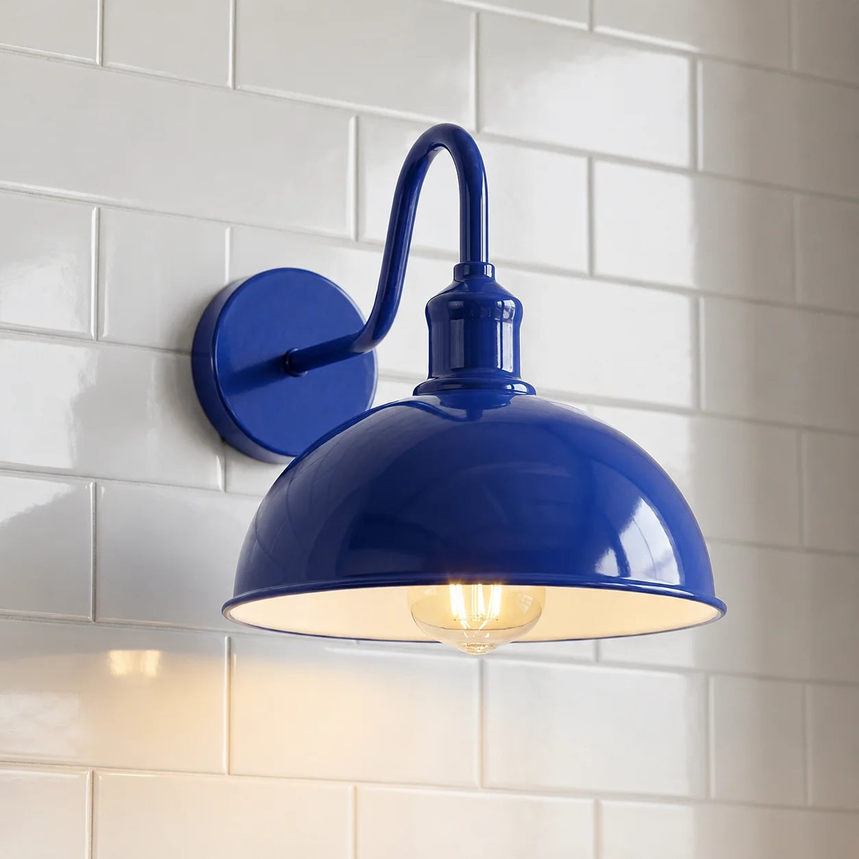 A glossy cobalt blue wall sconce with a curved gooseneck arm and a dome-shaped shade, shown illuminated against a white subway tile wall.