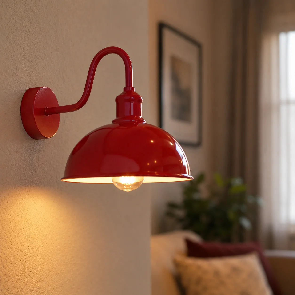 A red metal wall sconce with a curved arm and dome shade, shown lit on a beige wall in living room setting.