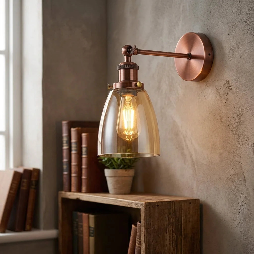 A copper-toned wall sconce with a clear glass bell shade and visible warm Edison bulb, mounted on a textured beige wall above a wooden shelf with books.