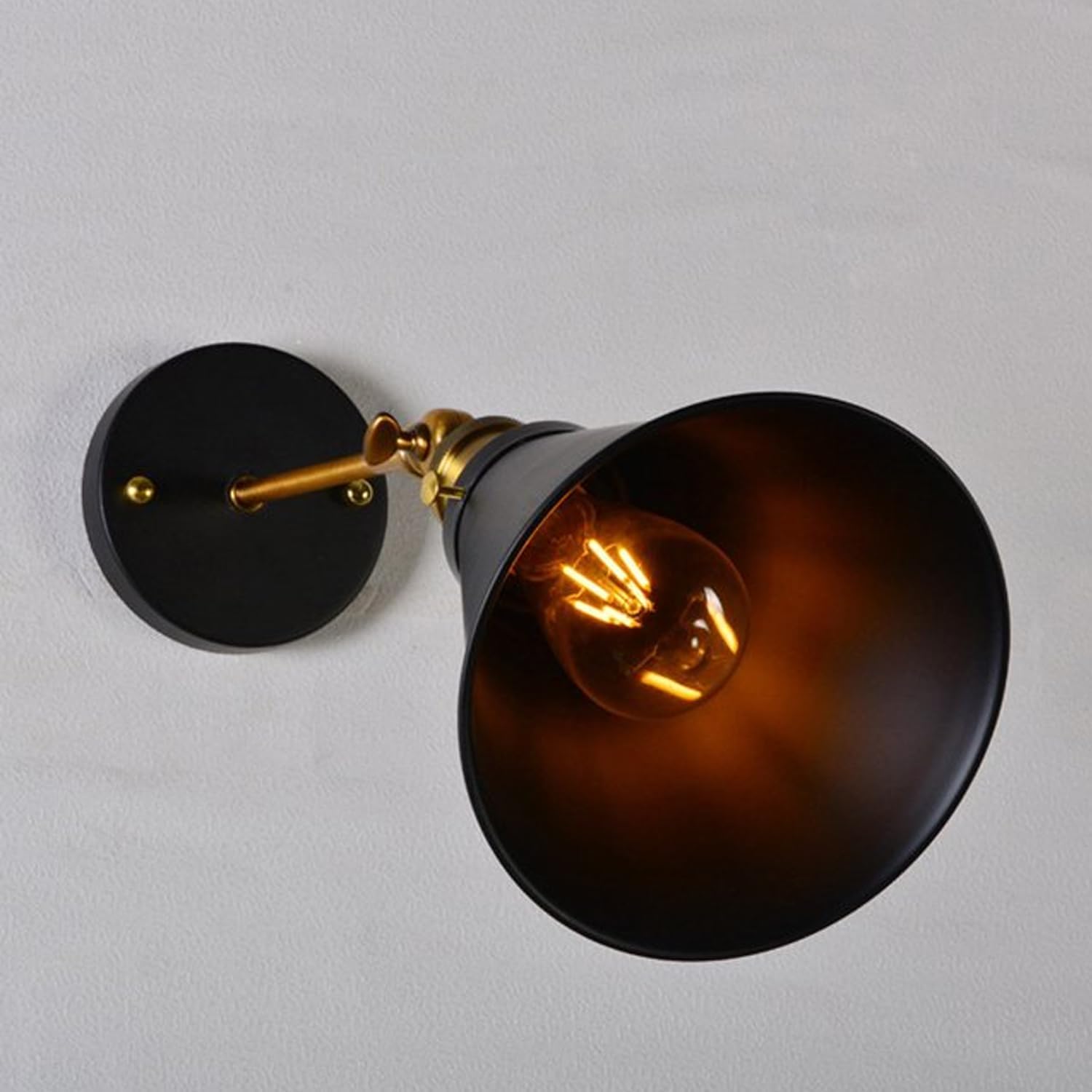 A close-up of an industrial black metal adjustable wall sconce with a gold-tone arm, featuring a cone-shaped shade and an illuminated bulb, mounted on a light-coloured wall.