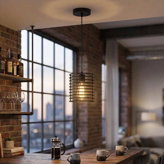 A modern industrial-style pendant light with a cylindrical metal cage and glowing bulb hangs above a rustic wooden bar counter in a loft apartment with brick walls, shelves, and bar stools.