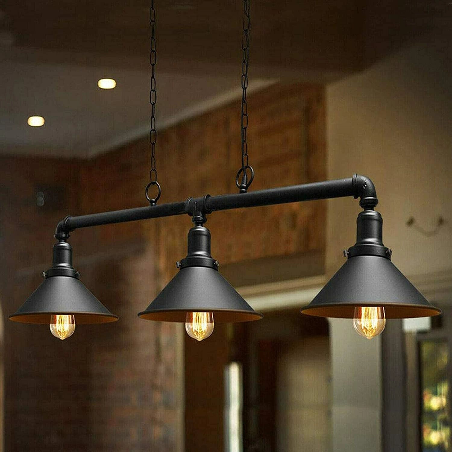 Three-light industrial pendant chandelier with black metal pipe-style arms and conical shades, shown hanging in a real interior with warm bulbs illuminated.