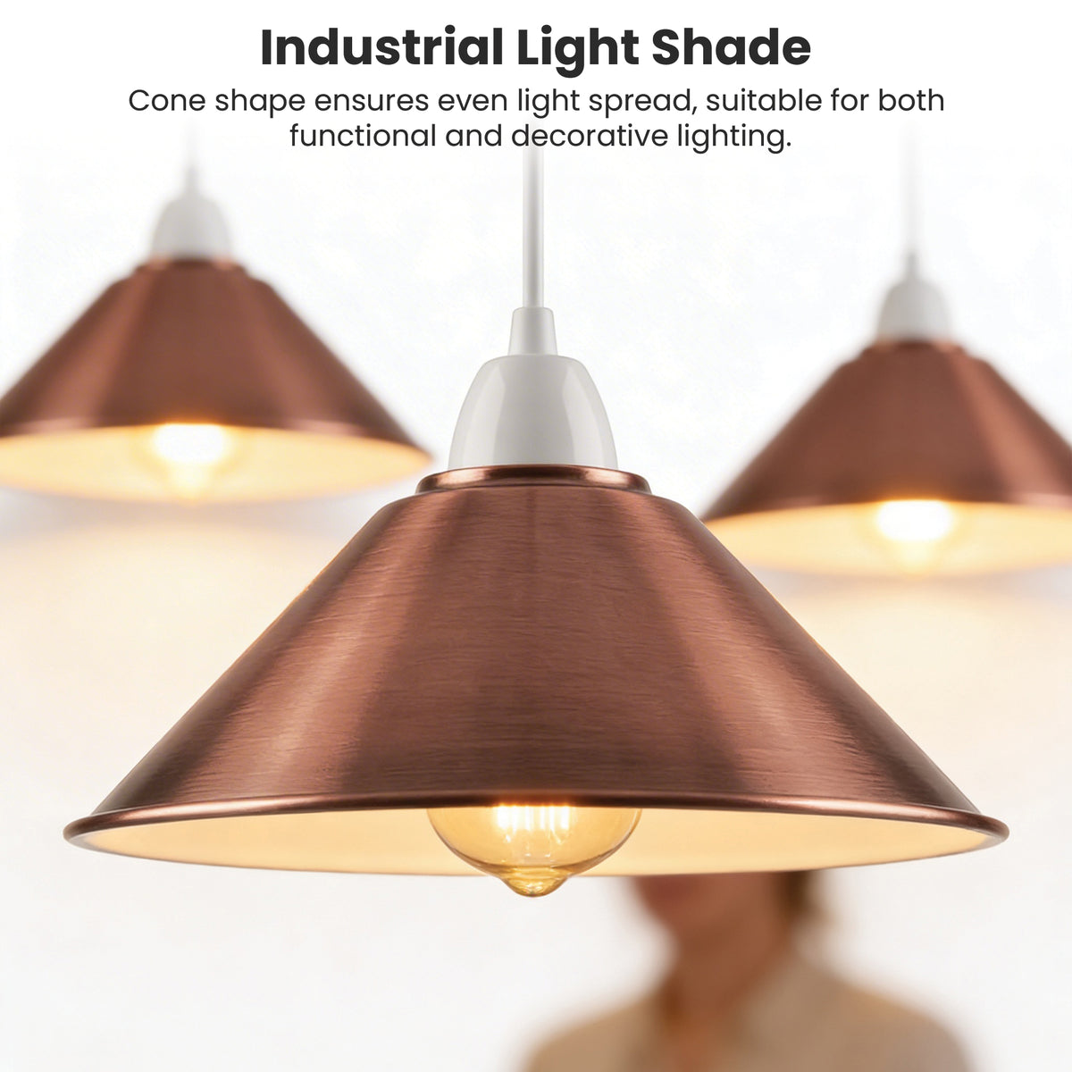 A copper-colored industrial pendant light shade with a white socket and glowing bulb, shown against a softly blurred background with two additional shades and text describing even light spread.