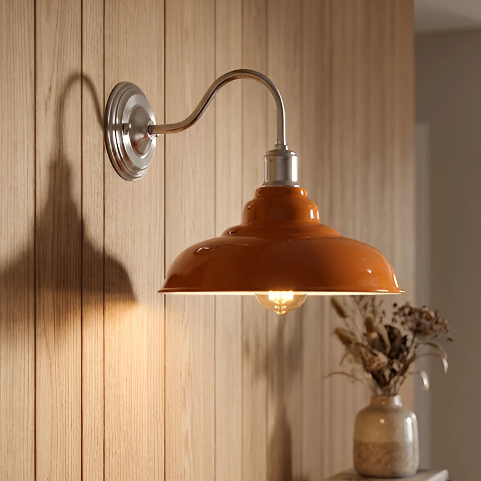 An industrial-style wall sconce with a glossy orange dome shade and a curved silver metal arm, mounted on a light wood-paneled wall. The warm light from the bulb illuminates the wall, and a small vase with dried stems sits on a surface below.