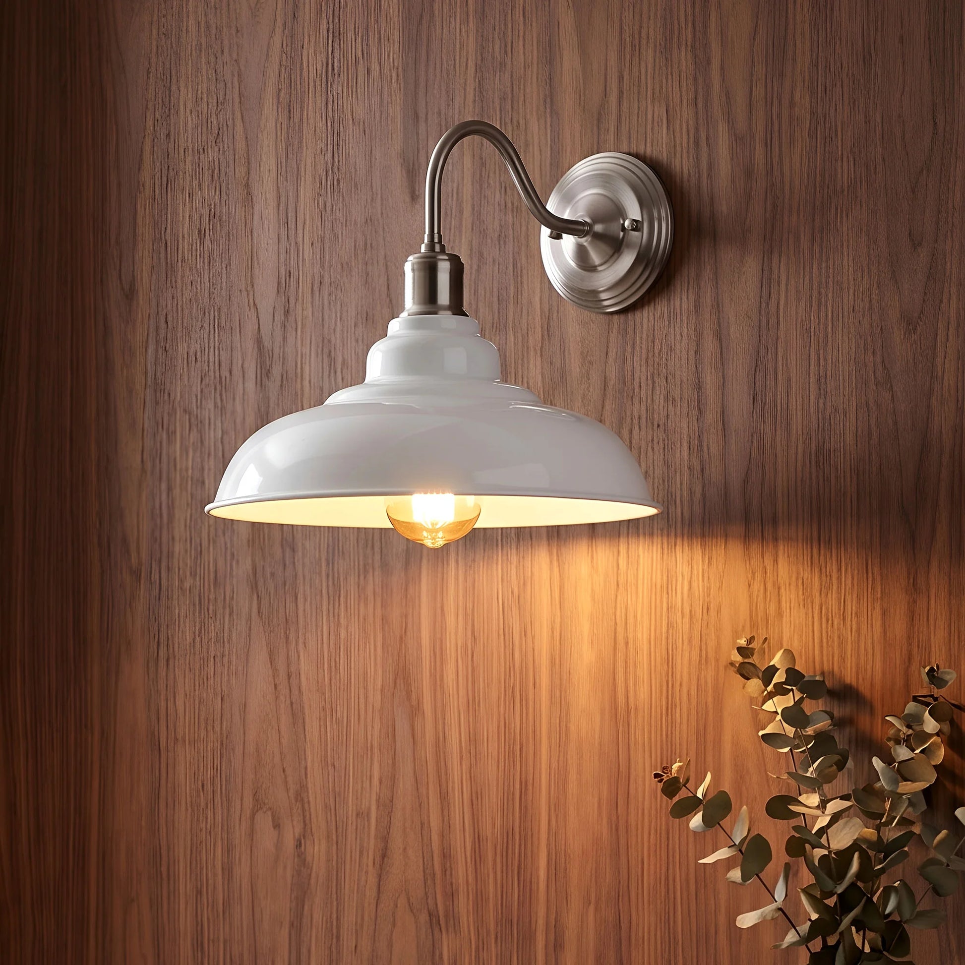 A wall-mounted sconce with a white dome-shaped glass shade and brushed metal arm and wall plate, shown lit against a brown wood-paneled wall with a small plant in the lower right corner.
