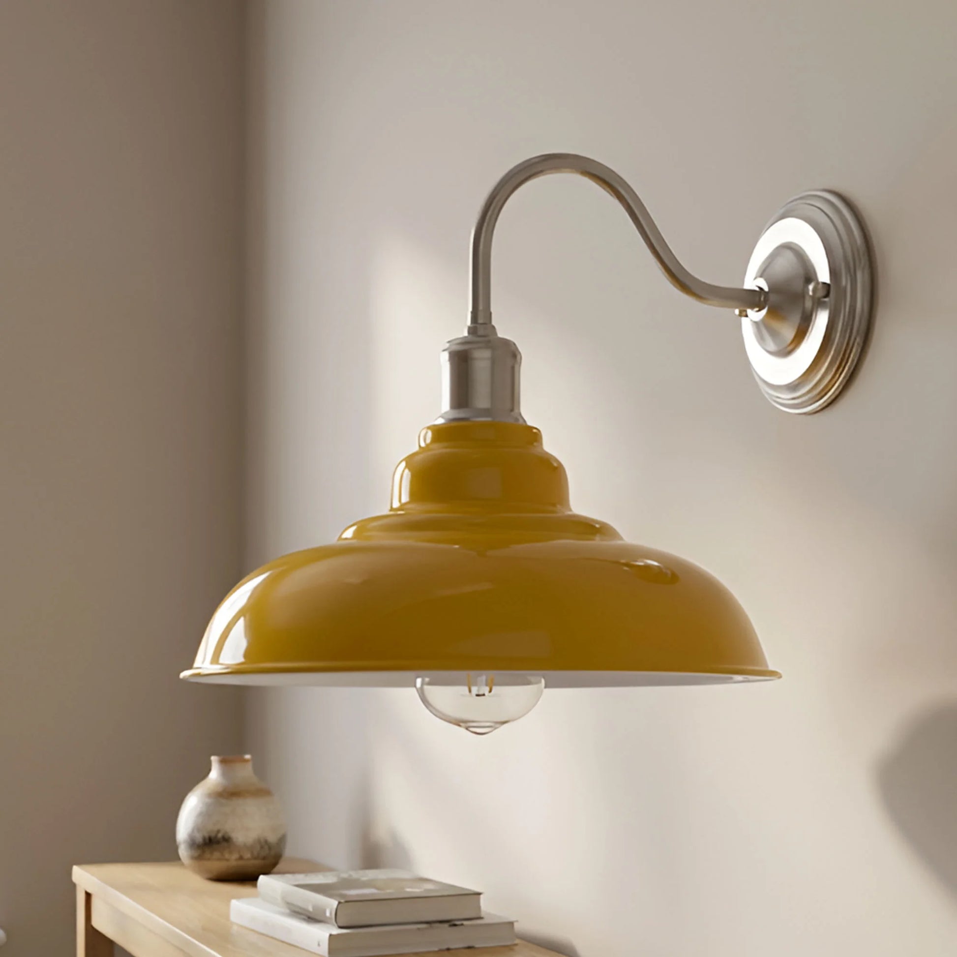 A wall-mounted sconce lamp with a glossy mustard yellow dome shade and a brushed silver metal arm and wall plate, shown lit against a warm beige wall above a small wooden table with books and a vase.