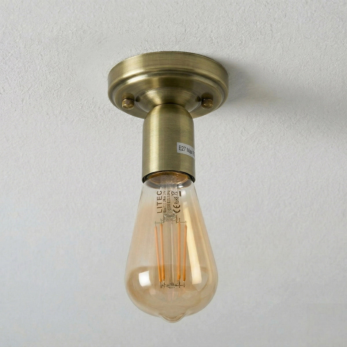 An industrial-style ceiling light fixture with a round brass ceiling mount and a clear glass bulb showing an Edison-style filament, mounted on a textured white ceiling.