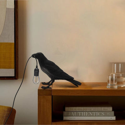 A black crow-shaped table lamp with a plug and a free E27 bulb, placed on a wooden table.