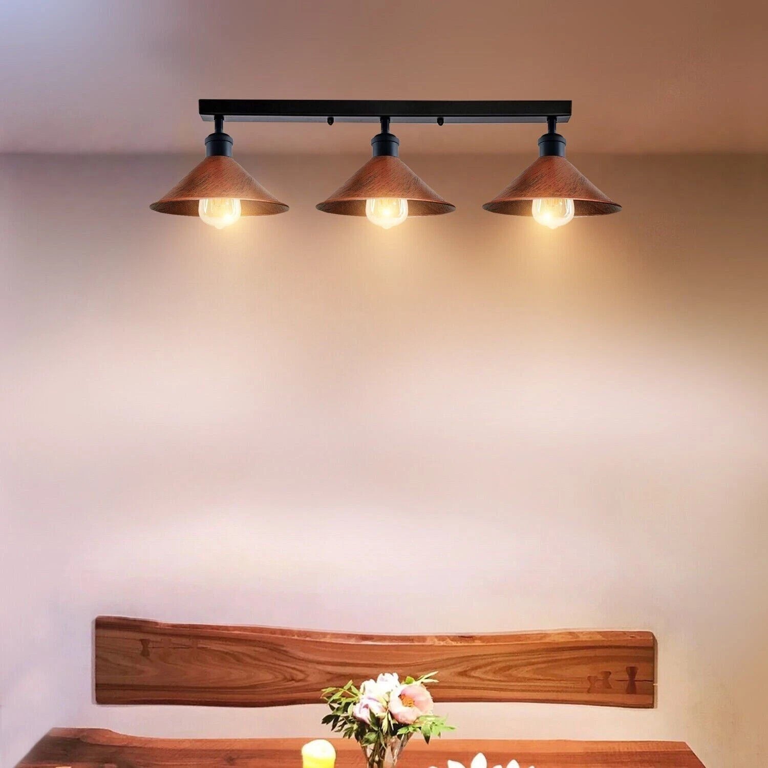 Three-light fixture on a wall above a wooden table with flowers.